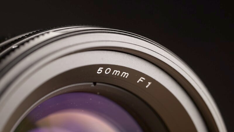 Close-up of a camera lens with "50mm F1" labeled on the outer ring. The lens has a metallic finish, and a portion of the glass is visible, reflecting various colors. The background is dark, emphasizing the details of the lens.