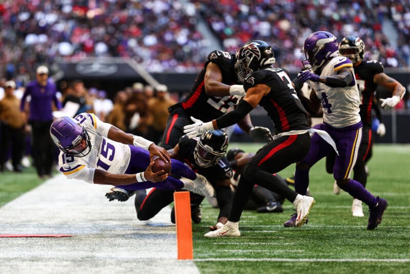 A football player in a white and purple uniform dives towards the end zone, extending the ball over the goal line. Three players in black and red uniforms try to stop him, while one teammate in a white and purple uniform looks on. The crowd watches the action.