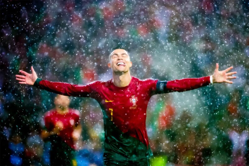 A soccer player stands in the rain with arms outstretched, eyes closed, and a joyful expression. He is wearing a red and green uniform with the number 7 and a captain's armband. The background is blurred, filled with raindrops and indistinct figures.