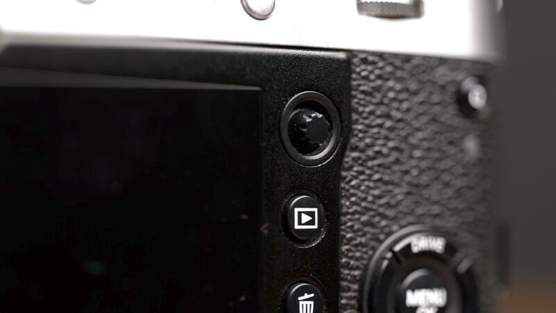 Close-up of the back of a camera, showing textured black and silver surfaces. Visible buttons include a playback button designated by a triangle icon and a delete button indicated by a trash can icon. The screen is partially visible on the left side.