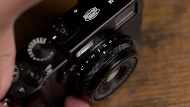 A close-up shot of a handheld camera, focusing on the lens and control dials. The camera features a manual aperture ring with visible f-stop numbers and textured metal detailing. The background is a wooden surface.