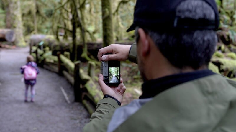 A person in a black baseball cap and green jacket is taking a picture with a camera in a mossy forest. The photo captures a child in a pink jacket and backpack walking ahead on a gravel path, surrounded by lush greenery and wooden railings.