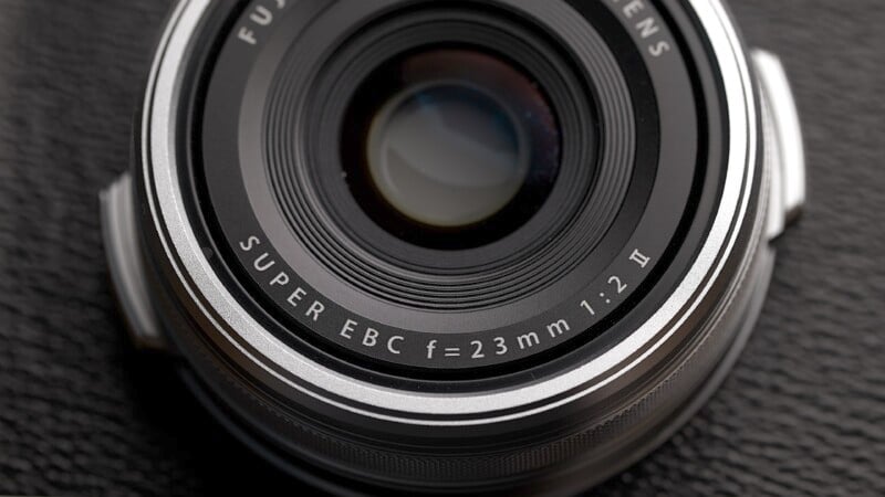 Close-up shot of a Fujifilm camera lens with the text "SUPER EBC f=23mm 1:2" engraved on the front rim. The lens is silver with a black interior and is resting on a textured black surface.