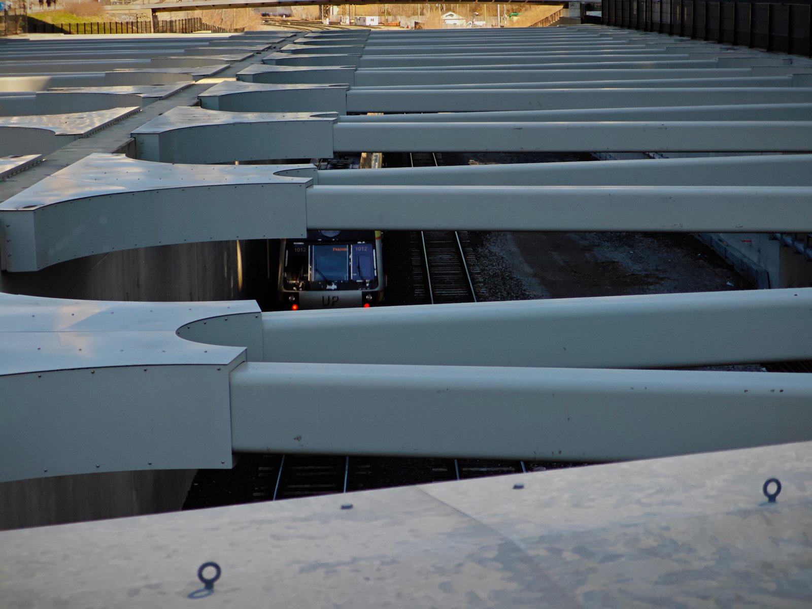 A train passing through a series of large, white beams structured in parallel rows, creating an architectural pattern, with a glimpse of tracks visible beneath.